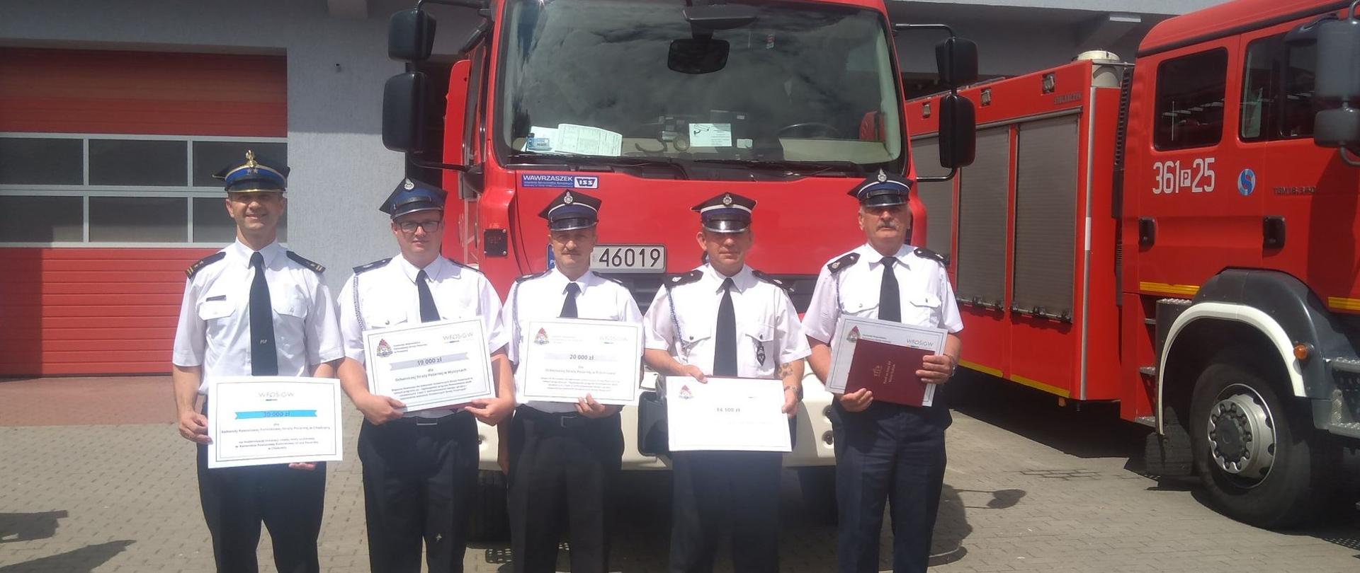 Zdjęcie przedstawia strażaków podczas uroczystego wręczenie promes dla jednostek Państwowej Straży Pożarnej i ochotniczych straży pożarnych.
W tle obiekty Komendy Miejskiej Państwowej Straży Pożarnej w Koninie.
