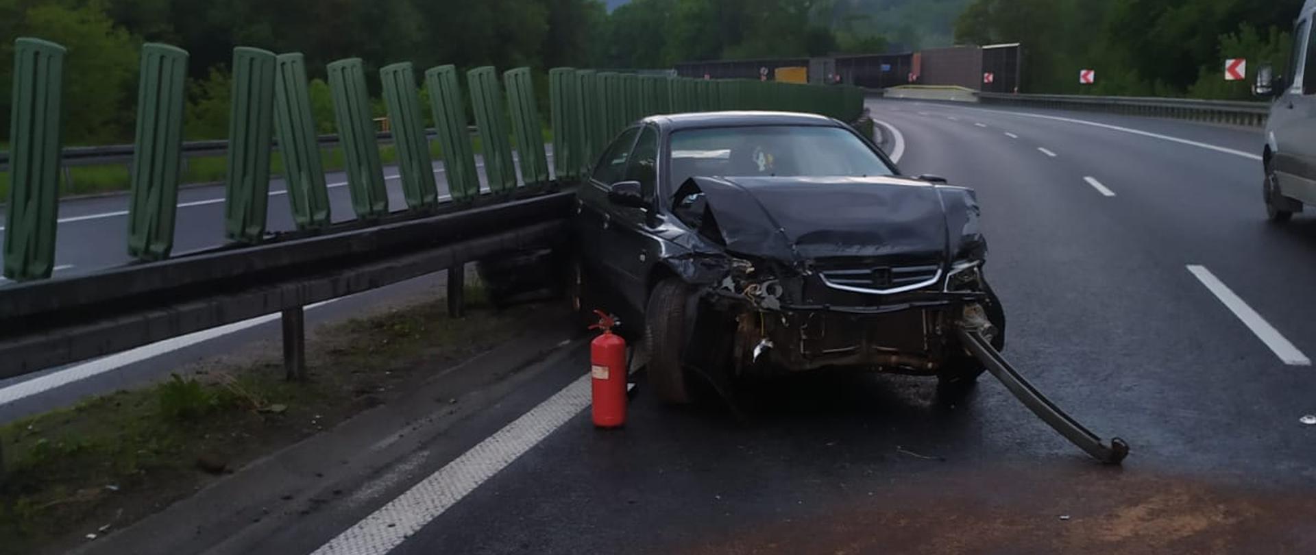 Na zdjęciu widoczne są pasy ruchu na drodze ekspresowej. W centralnej części stoi rozbity samochód osobowy w kolorze czarnym. Przy nim położona jest gaśnica strażacka, a na jezdni rozsypany sorbent pokrywający substancje ropopochodną.