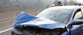 Na zdjęciu widoczny niebieski samochód osobowy z rozbitym przodem po uderzeniu w bariery ochronne. Za nim częściowo widoczni funkcjonariusze policji i straży pożarnej wykonujący czynności służbowe.