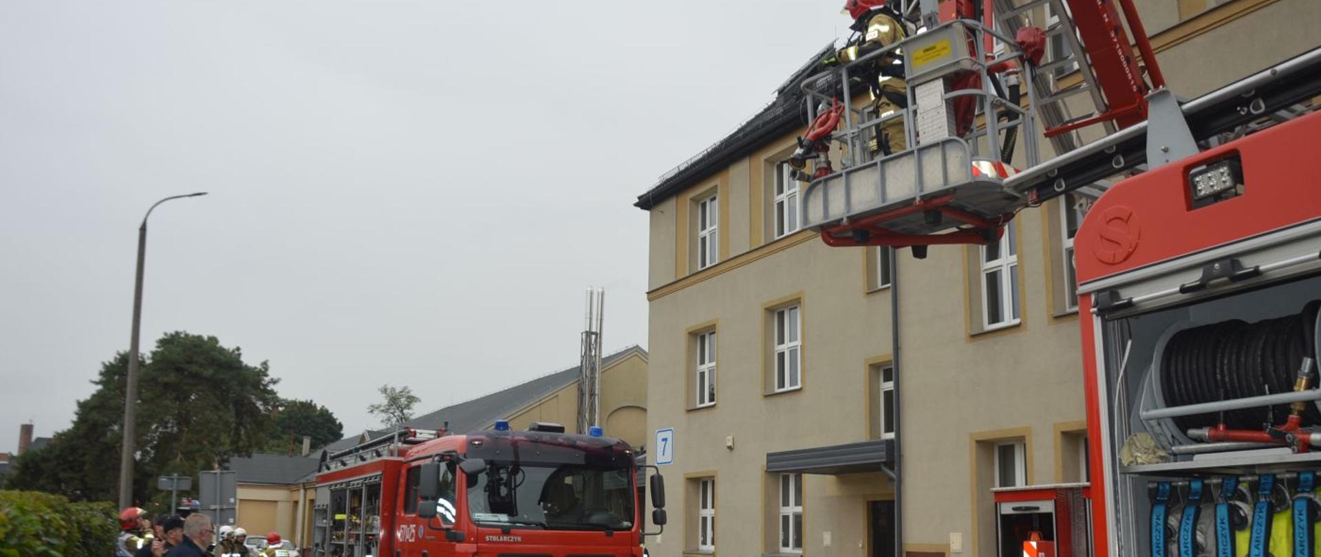 Ćwiczenia na terenie szpitala. Teren przed Zakładem Opiekuńczo-Leczniczym. Na drodze pożarowej stoją trzy samochody pożarnicze, w tym widoczny jest kosz podnośnika hydraulicznego, a w nim strażak. Wozy strażackie mają otwarte skrytki ze sprzętem. W tle drzewa, obserwatorzy ćwiczeń oraz strażacy biorący w nich udział.