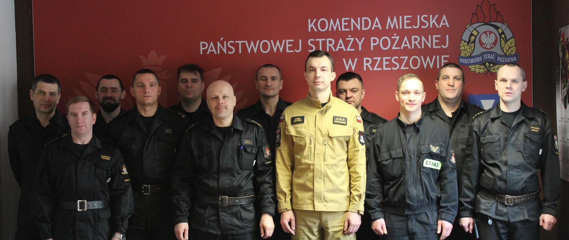 Zdjęcie przedstawia funkcjonariuszy PSP w umundurowaniu. Są ustawieni w sali szkoleniowej w dwóch rzędach i umundurowani czarne umundurowanie dowódczo-sztabowe. Wyjątek stanowi funkcjonariusz umundurowany w piaskowe umundurowanie służbowe wprowadzana obecnie w PSP oraz drugi funkcjonariusz umundurowany w ubranie koszarowe wycofywane obecnie w PSP. Wszyscy znajdują się na tle ścinki z napisem Komenda Miejska Państwowej Straży Pożarnej w Rzeszowie.