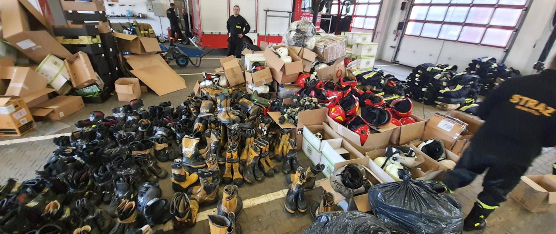 na zdjęciu dwóch strażaków oraz bardzo duże ilości butów hełmów i ubrań strażackich w tle samochód gaśniczych, zdjęcie zrobione w garażu jednostki ratowniczo gaśniczej