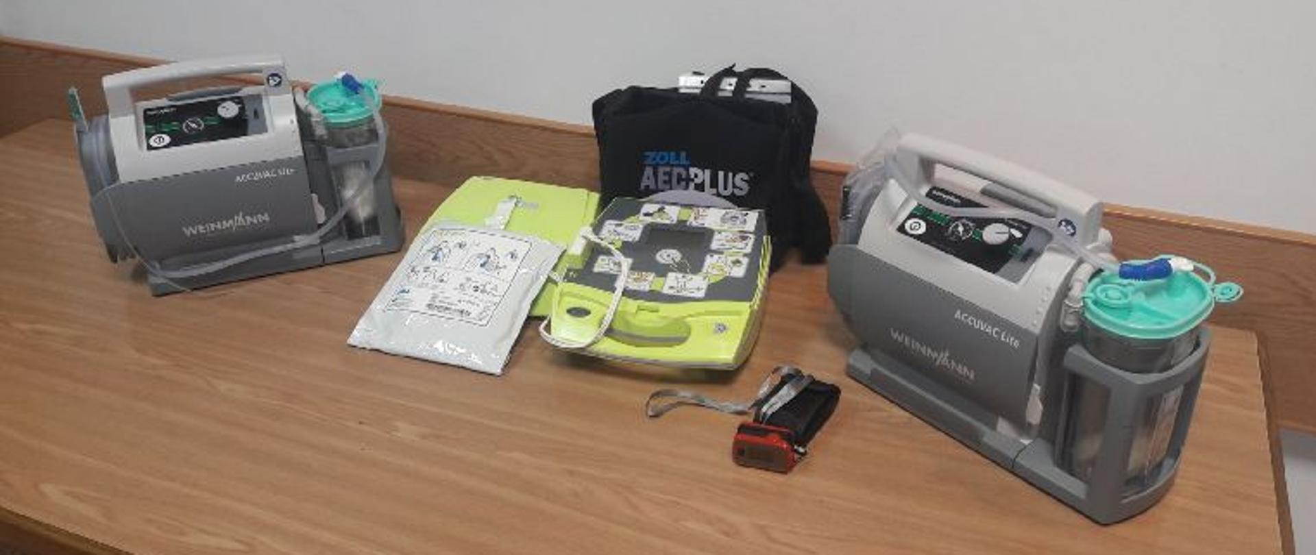 Zdjęcie przedstawia ułożony na brązowym stole na tle białej ściany, nowy sprzęt medyczny w Jednostce Ratowniczo-Gaśniczej Komendy Powiatowej Państwowej Straży Pożarnej w Brzozowie. Po lewej i po prawej stronie stoją dwa takie same szaro-popielate prostokątne urządzenia (ssaki akumulatorowe) z uchwytami. Na środku fotografii zielono-szary zautomatyzowany defibrylator (AED) elektrodami, obok czarny pokrowiec na defibrylator. Z przodu leży małe czerwone urządzenie elektroniczne pulsoksymetr w formie klipsu.