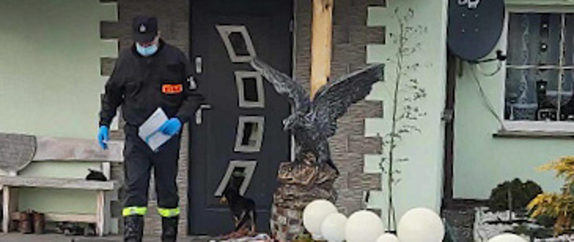 Na zdjęciu znajduje się strażak, który roznosi ulotki. Ubrany jest w ubranie koszarowe, a w lewej ręce ma ulotki. Za strażakiem znajduje się zielony dom, a sam strażak jest na podwórku. Zdjęcie wykonano w dzień.