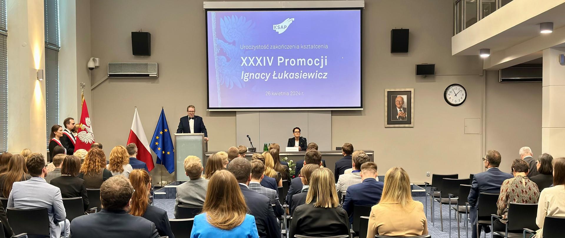 Aula pełna ludzi, po lewej stronie poczet sztandarowy, na środku przy mównicy dyrektor KSAP, obok szefowa służby cywilnej.