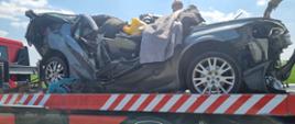 Tragiczny wypadek. Wrak rozbitego samochodu znajduje się na autolawecie. Za nim widoczna głowa strażaka w czerwonym hełmie.