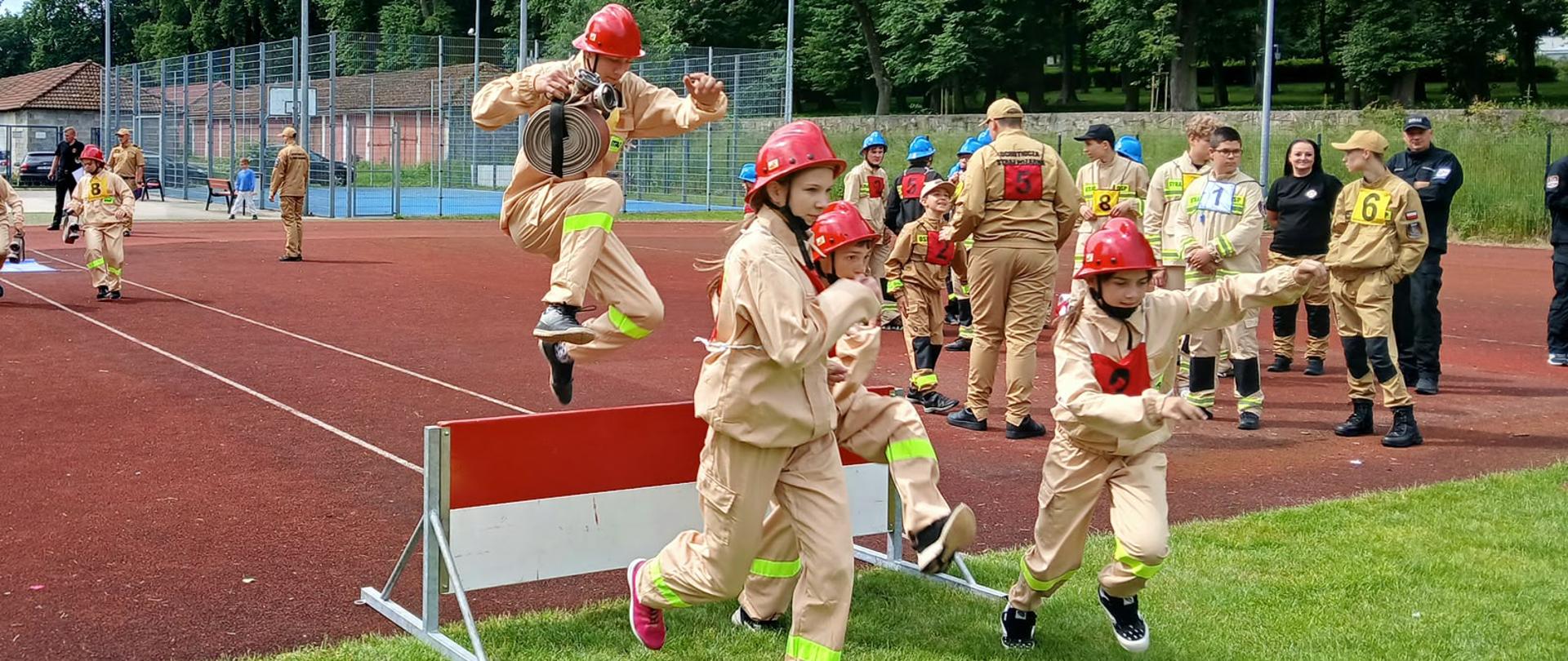 Młodzieżowe zawody pożarnicze na stadionie lekkoatletycznym. Na pierwszym planie chłopak w piaskowym mundurze młodzieżowej drużyny pożarniczej i czerwonym hełmie przeskakuje w biegu przez biało-czerwoną przeszkodę, trzymając w rękach zwinięty wąż strażacki. Obok niego, przez przeszkodę i obok niej, dynamicznie przebiegają inne dzieci w podobnych strojach. W tle widać pozostałych uczestników zawodów, zielone drzewa oraz fragment bocznego boiska.
