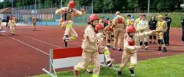 Młodzieżowe zawody pożarnicze na stadionie lekkoatletycznym. Na pierwszym planie chłopak w piaskowym mundurze młodzieżowej drużyny pożarniczej i czerwonym hełmie przeskakuje w biegu przez biało-czerwoną przeszkodę, trzymając w rękach zwinięty wąż strażacki. Obok niego, przez przeszkodę i obok niej, dynamicznie przebiegają inne dzieci w podobnych strojach. W tle widać pozostałych uczestników zawodów, zielone drzewa oraz fragment bocznego boiska.