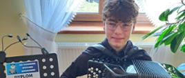 Zdjęcie kolorowe – Stanisław Rogala siedzi na krześle i trzyma na kolanach akordeon, ubrany jest w czarną koszulę i czarne spodnie, a obok na pulpicie stoi dyplom za I miejsca w XXX Kaszubskich Spotkaniach Akordeonowych.
