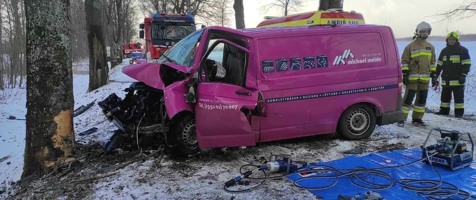 Na zdjęciu rozbity o drzewo samochód bus ustawiony w poprzek drogi. Przy samochodzie strażacy i na drodze niebieska plandeka na której poukładane narzędzia hydrauliczne. W tle samochody strażackie.