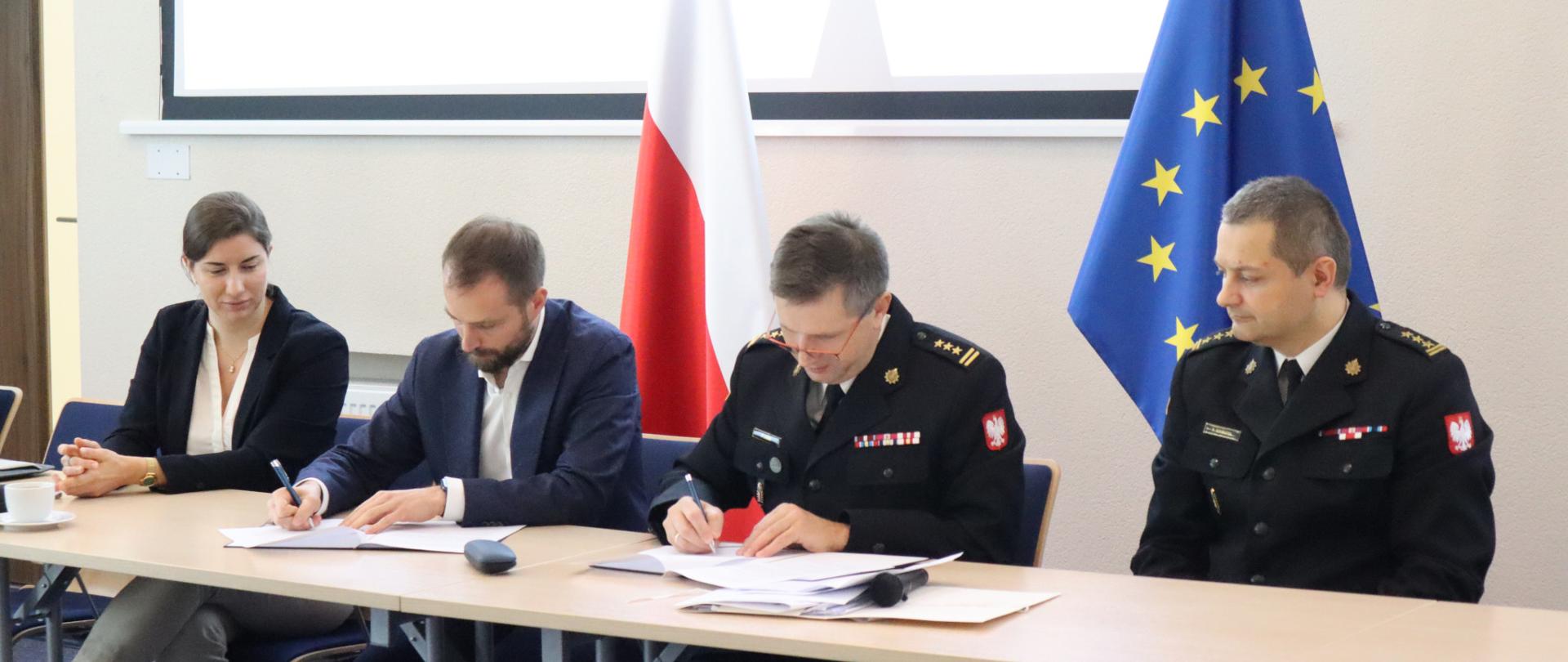 Na zdjęciu znajdują się przedstawiciele PSPA, dyrektor CNBOP-PIB oraz zastępca dyrektora Biura Przeciwdziałania Zagrożeniom KG PSP podczas podpisywania porozumienia przy stole konferencyjnym. W tle flaga Polski i UE 