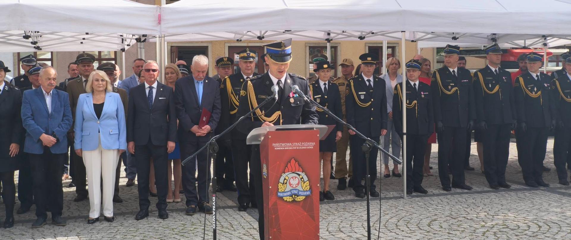 Oficjalne uroczystości Państwowej Straży Pożarnej na brukowanym placu miejskim. Na pierwszym planie za mównicą z godłem strażackim stoi przemawiający funkcjonariusz w galowym mundurze i czapce garnizonowej. Za nim, pod ciągiem połączonych białych namiotów, stoją zaproszeni goście w garniturach i garsonkach oraz liczny szpaler strażaków w pełnym umundurowaniu wyjściowym. W tle widoczne są ściany zabytkowych kamienic oraz ramię wysuniętej drabiny mechanicznej wozu strażackiego.