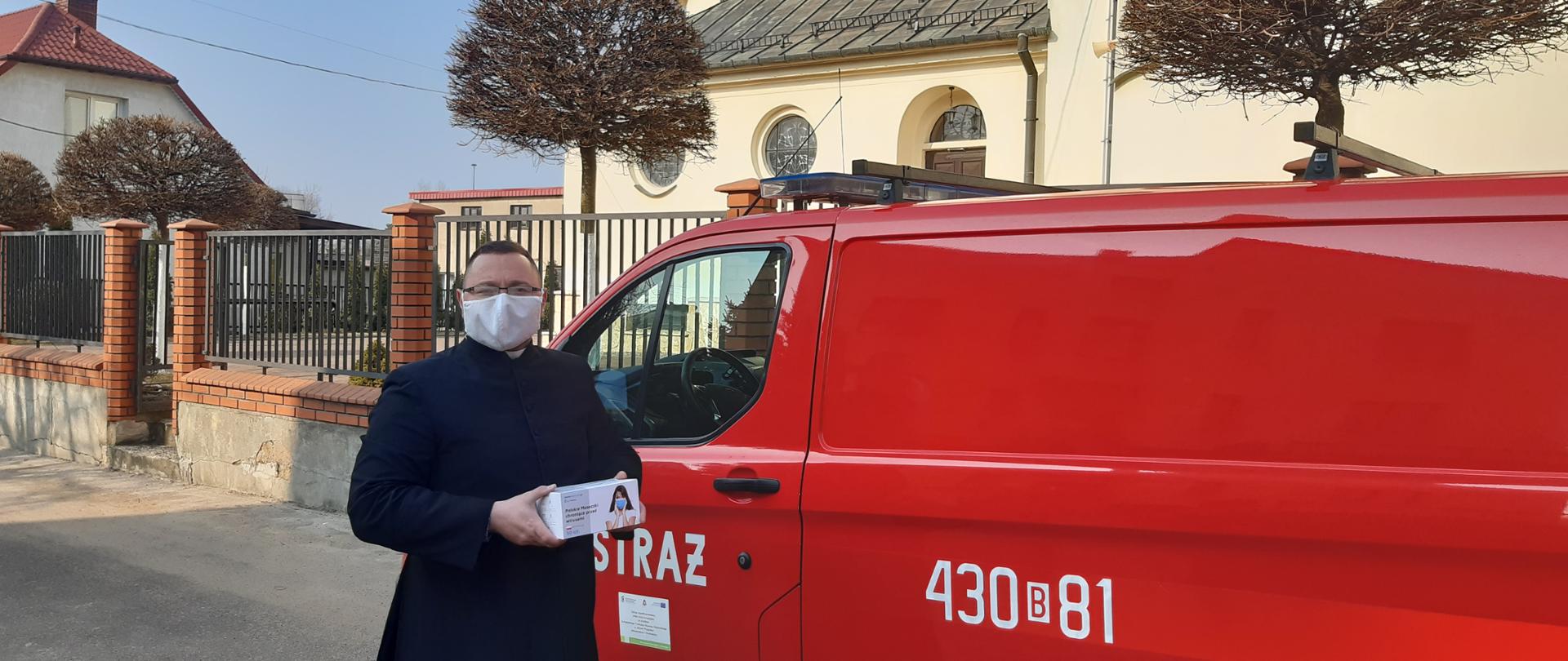 Na tle czerwonego strażackiego busa na pierwszym planie stoi ksiądz w sutannie trzymający pudełko z maseczkami. Za samochodem żółty kościół.