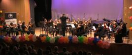 Zdjęcie kolorowe. Orkiestra na scenie podczas występu w sali koncertowej. Dyrygent tyłem do widowni, z uniesionymi rękami, kieruje grupą muzyków grających na skrzypcach, wiolonczelach i kontrabasach. Muzycy ubranina czarno lub w stroje galowe, siedzą na krzesłach przed pulpitami z nutami. Krawędź sceny udekorowana rzędem kolorowych balonów. W tle duży jasny ekran oświetlony fioletowym światłem oraz drewniane panele ścienne. Po lewej stronie baner szkoły muzycznej. Na pierwszym planie ciemne sylwetki publiczności siedzącej w rzędach foteli.