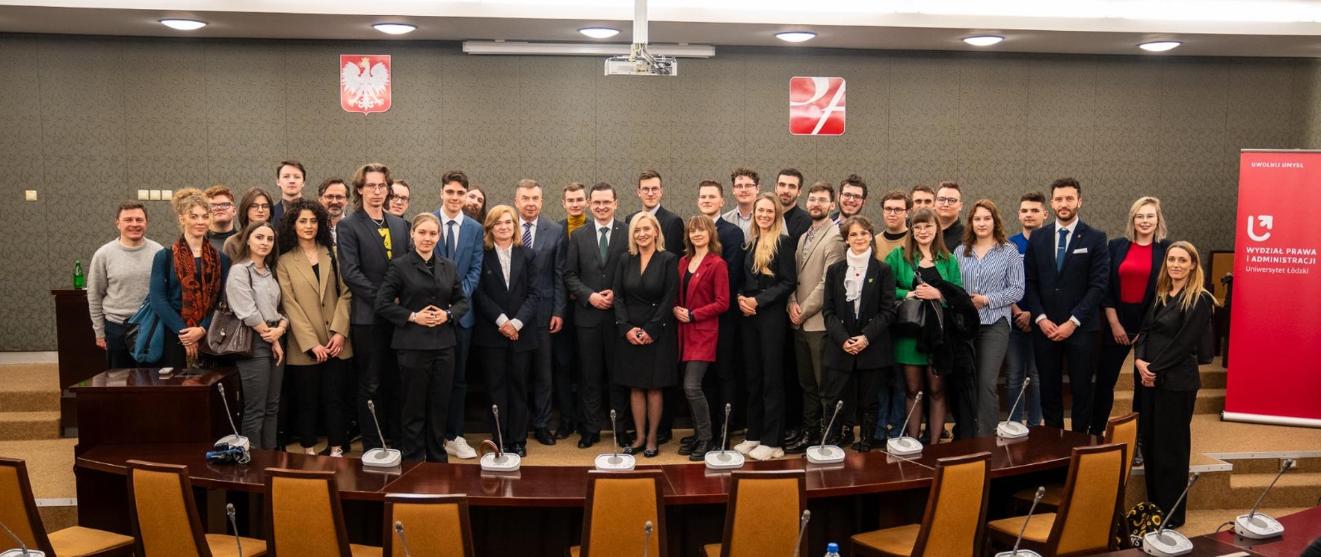 Zdjęcie zbiorowe, pośrodku dużej sali pod ścianą stoi duża grypa ludzi, przed nimi ustawione w łuk stoły i krzesła.