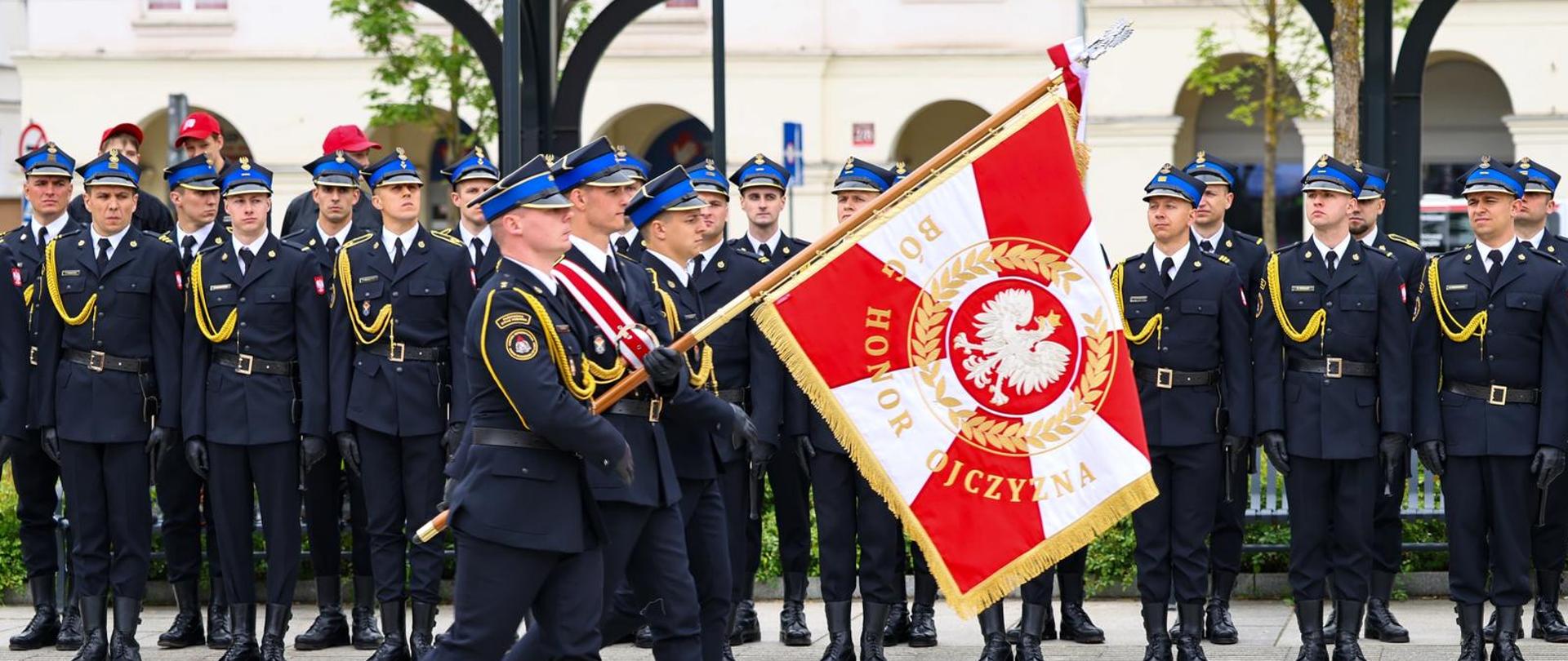 Poczet sztandarowy PSP maszeruje ze sztandarem z hasłem „Bóg, Honor, Ojczyzna”. W tle stoi pododdział honorowy.