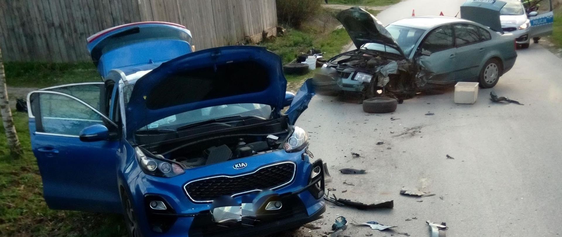 Na pierwszym planie zdjęcia niebieski samochód osobowy z otwartymi drzwiami i przednią maską z uszkodzonym przodem, wokół pojazdu porozrzucane elementy karoserii, w dalszym planie drugi samochód osobowy z otwartą maską i leżącym obok oderwanym kołem przednim. W tle za samochodami widoczna drewniany budynek gospodarczy, przejazd na drodze zablokowany przez stojące na drodze pojazdy.