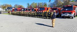 Na zdjęciu widać zbiórkę strażaków na placu przed jednostką. Ustawieni są w szeregu, w pełnym umundurowaniu bojowym, a za nimi stoi rząd czerwonych wozów strażackich.
Na pierwszym planie znajduje się dowódca, który stoi naprzeciwko grupy i prawdopodobnie wydaje polecenia albo prowadzi odprawę.