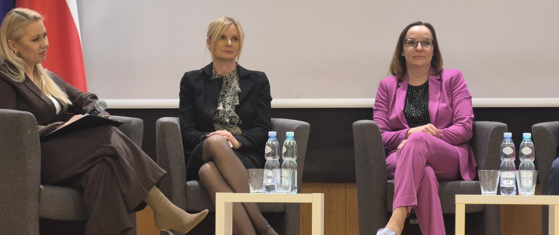 Zdjęcie przedstawia scenę z panelu dyskusyjnego, trzy kobiety siedzą w fotelach ustawionych w rzędzie. Kobieta w środku, blondynka, trzyma mikrofon
i mówi do niego. W tle, za uczestniczkami, widać dwie flagi: jedną z gwiazdami na niebieskim tle oraz drugą w biało czerwonych – barwach