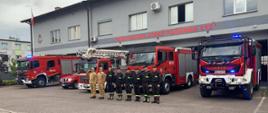 Strażacy w mundurach stoją w szeregu przed pojazdami strażackimi i budynkiem remizy. Pierwszy od lewej strony strażak w szeregu, oddaje honor salutując. 