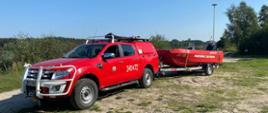 Na zdjęciu pojazd JOP w kolorze czerwonym z podczepioną przyczepą do łodzi, na której znajduje się czerwona łódź. Zdjęcie w ciągu dnia. Słonecznie. Podłoże piaszczyste.