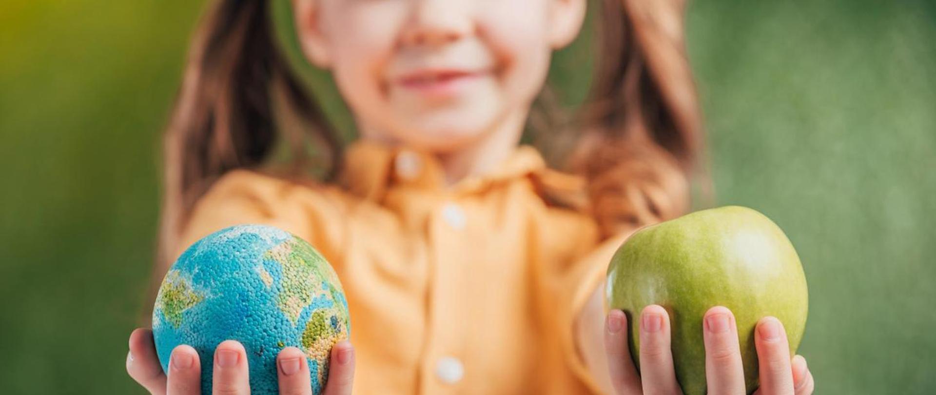 Zdjęcie przedstawia dziewczynkę trzymającą w prawej ręce globus a w lewej jabłko. 