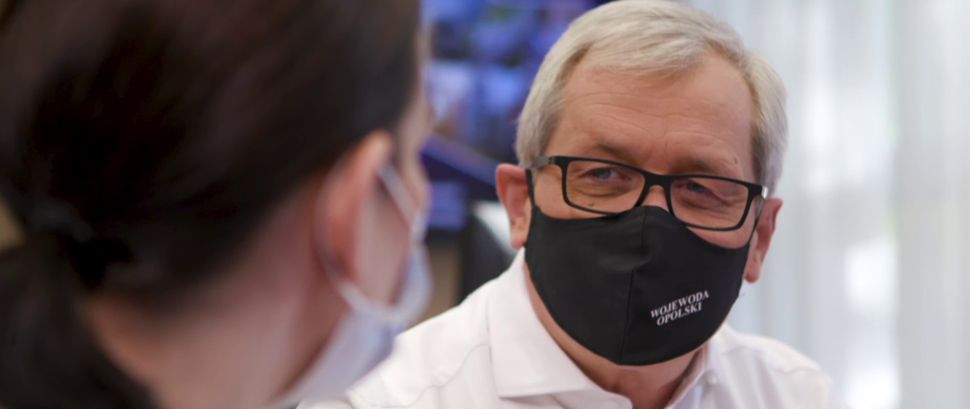 Zdjęcie - stop klatka TVP3 Opole. Przedstawia wojewodę opolskiego, prowadzącego rozmowę z pracownikiem urzędu wojewódzkiego w Opolu. Wojewoda znajduje się w centralnej części kadru, po lewej stronie pracownik ustawiony tyłem do obiektywu. Wojewoda ubrany jest w białą koszulę, czarną maseczkę ochronną. Na twarzy ma okulary korekcyjne, spogląda na pracownika. W tle monitor telewizyjny i okno. Bliski kadr.