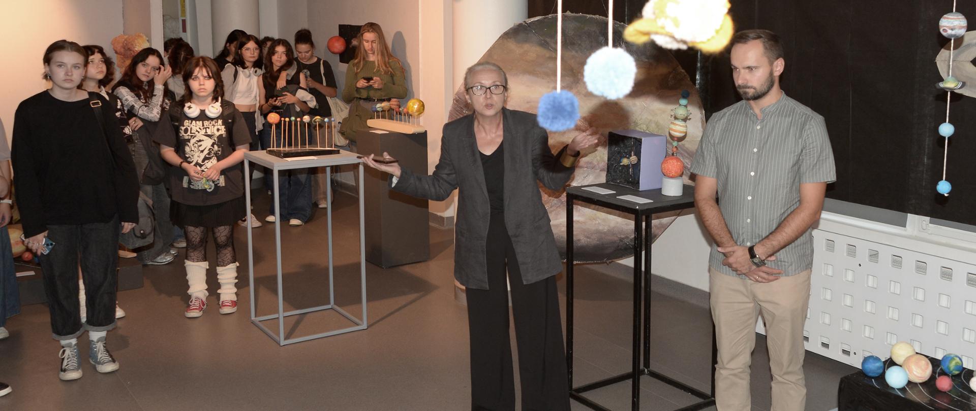 W galerii PLSP w Lublinie ściany i kolumny pomalowane są na biało. Podłoga jest szara. We wnętrzu znajdują się ściany z białych płyt. Okna są przyciemnione przez zasłonięcie ich czarnym materiałem. Na wystawie znajdują się modele układów słonecznych w różnych kompozycjach, planety, orbity, inne obiekty kosmiczne, itd. Uczniowie stoją i słuchają wice dyr Joanny Kozłowskiej, która mówi i ma rozłożone ręce. Obok niej stoi Tomasz Banaszkiewicz.