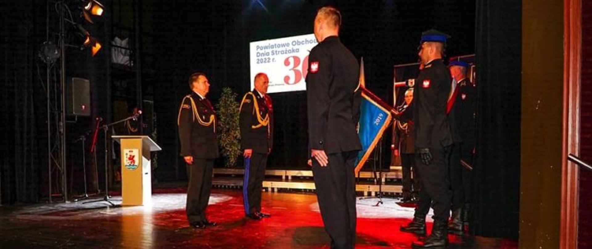 Widok z boku na scenę. Na scenie Komendant Powiatowy i Wojewódzki. Przed nimi poczet sztandarowy.