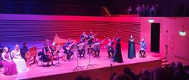 Grający zespół instrumentalny (9 muzyków), dwie wokalistki i wokalista siedzący na krzesłach, dwie stojące wokalistki w czarnych płaszczach na scenie sali koncertowej PSM