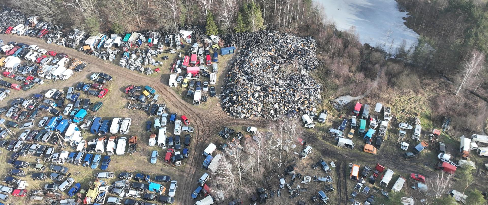 Widok z góry na kilkadziesiąt pojazdów i hałdy wymontowanych części obok lasu i stawu 