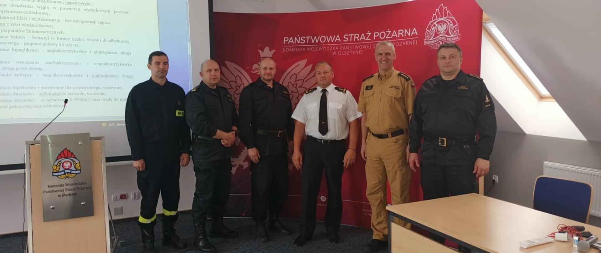 sześciu mężczyzn na czerwonym tle, po prawej stronie stoi drewniane biurko