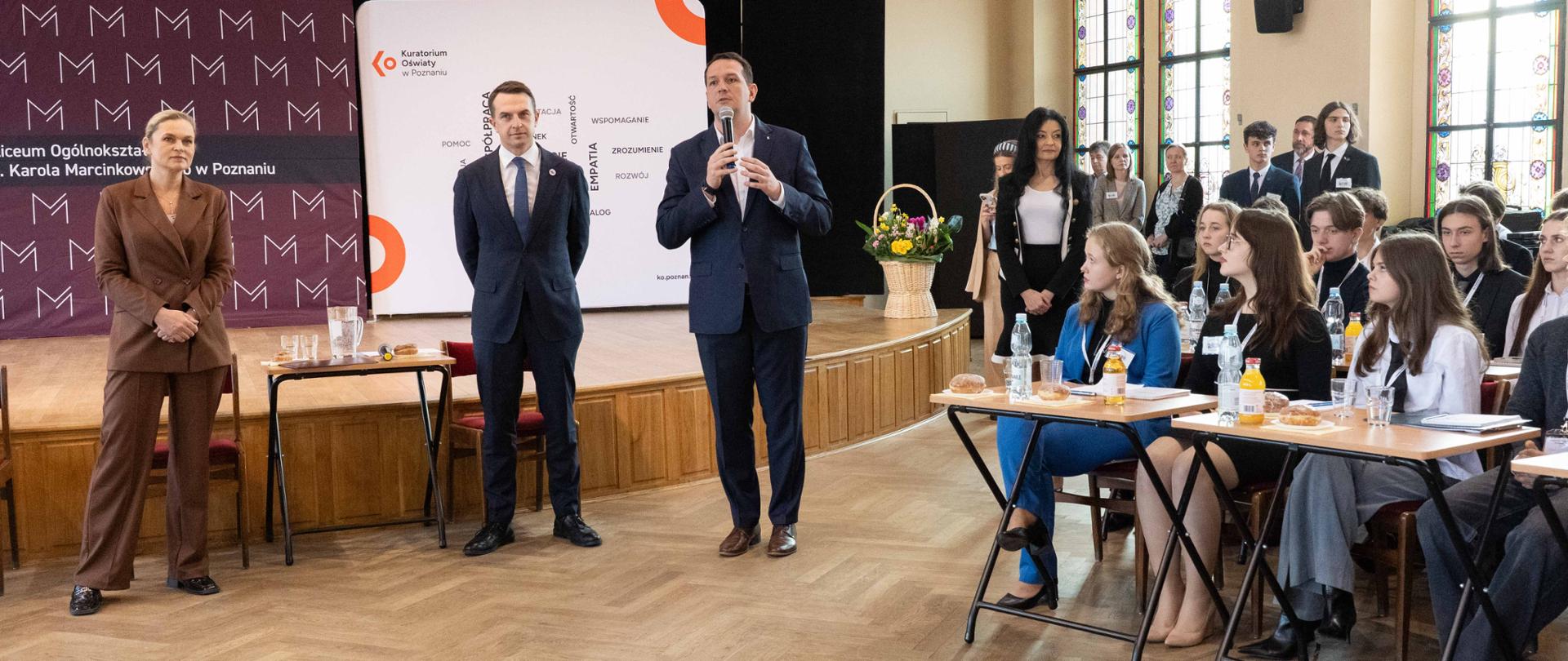 wizyta minister Edukacji w Poznaniu Na zdjęciu przed sceną widać stojącą minister Barbarę Nowacką. Po prawej stronie minister Szłapka oraz kurator oświaty. z prawej strony na krzesłach i przy stolikach siedzi młodzież. 