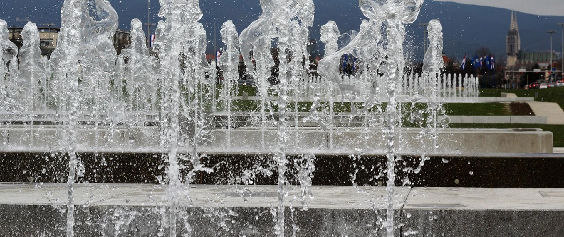 zdjęcie przedstawia fontannę, na pierwszym planie tryskające cztery strumienie wody, na drugim planie kolejne pięć strumieni wody, w oddali góry oraz zachmurzone niebo