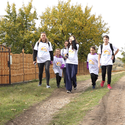 Kobieta i pięć dziewczynek idą wiejską drogą mijając zabudowania z drewnianym płotem