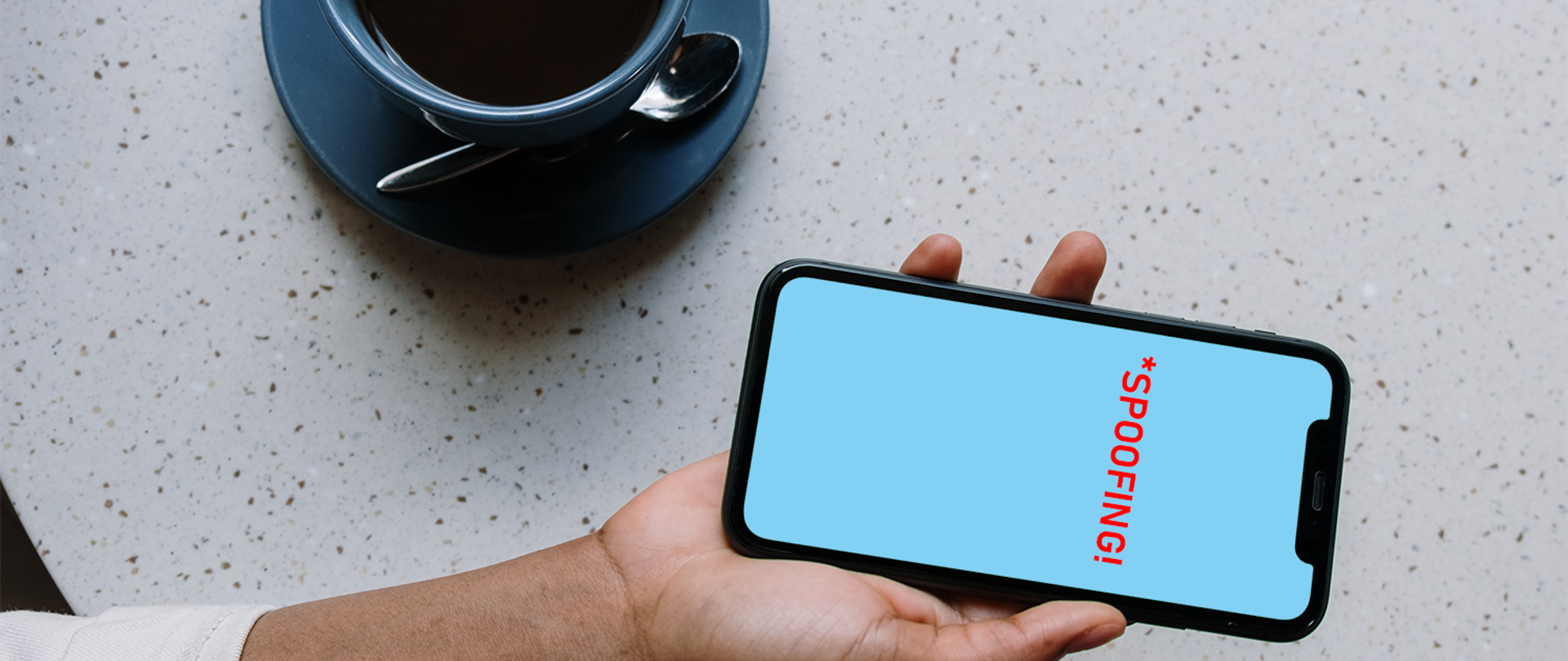 Zdjęcie przedstawia dłoń trzymającą smartfon obok stojącej filiżanki z kawą. Na ekranie smartfonu widoczny napis SPOOFING w kolorze czerwonym.