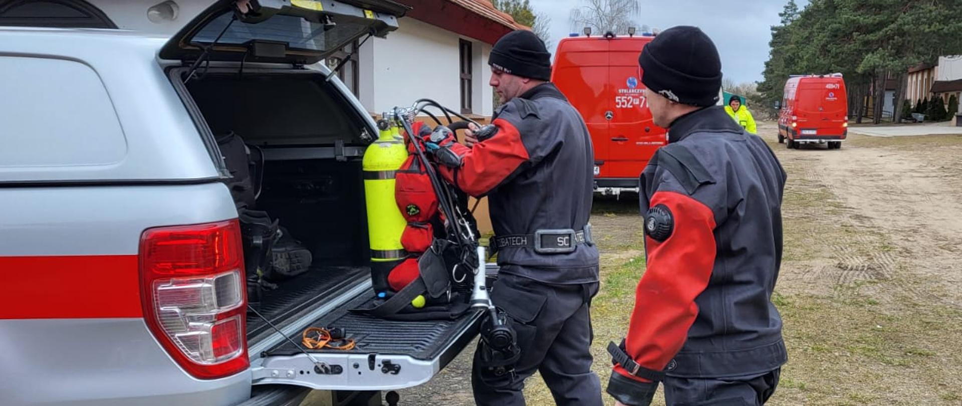 mężczyzna wyciąga z samochodu butlę do nurkowania, za nim stoi drugi mężczyzna, w tle samochody strażackie
