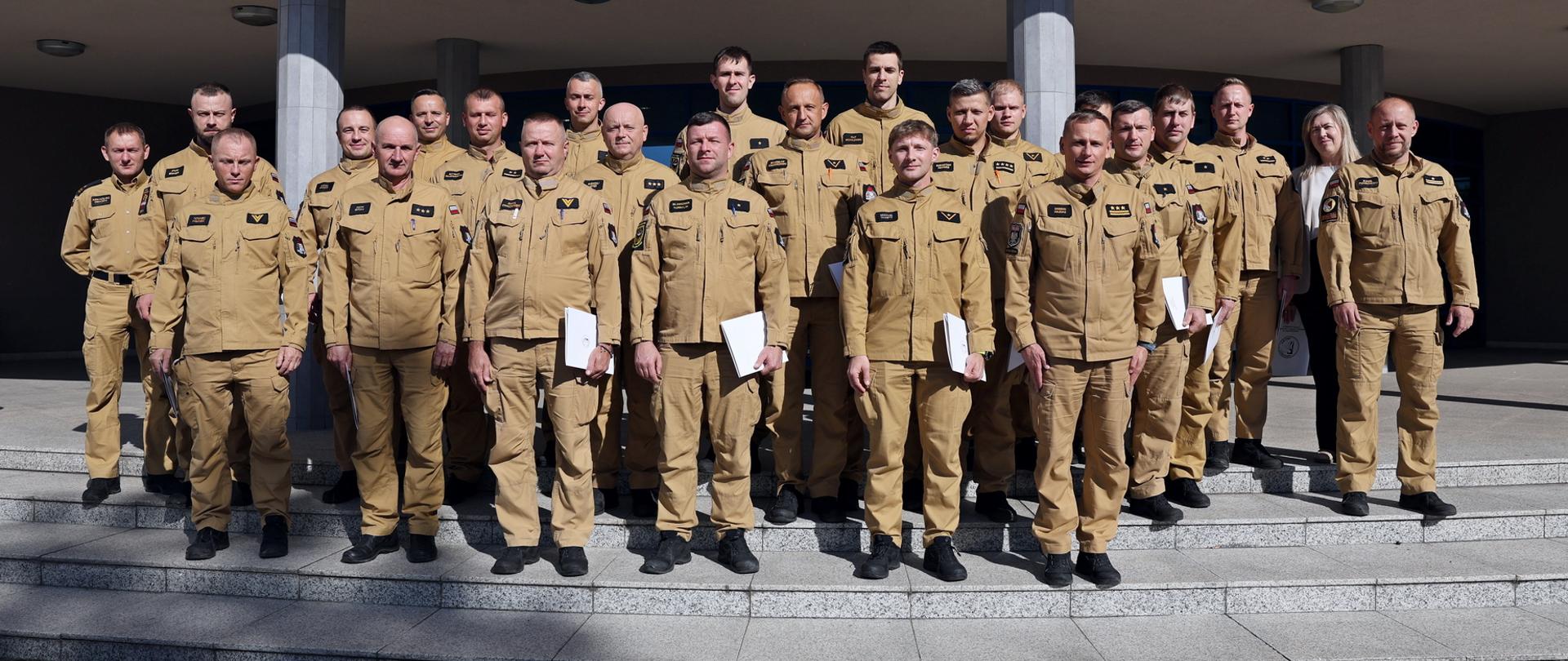 Pamiątkowe zdjęcie grupowe funkcjonariuszy PSP będących uczestnikami szkolenia z zakresu kierowania działaniem ratowniczym na poziomie interwencyjnym (20 osób) w towarzystwie Zastępcy Komendanta CS PSP, kobiety oraz dwóch funkcjonariuszy CS PSP