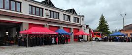 Widok szeroki na plac, na którym znajdują się namioty, pod którymi stoją uczestnicy uroczystości, czerwony wóz strażacki. Przy placu widoczny duży kremowy budynek. Powyżej pochmurne niebo.