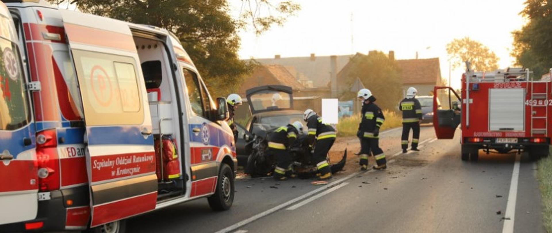 Drogę całkowicie blokują pojazdy służb ratunkowych, ponieważ na jednym pasie stoi karetka pogotowia, natomiast drugi pas blokuje samochód strażacki. Przed karetką widoczny jest rozbity samochód osobowy z mocno zdeformowanym przodem. Strażacy w białych hełmach i ubraniach bojowych wykonują dostęp do komory silnika samochodu osobowego celem odłączenia akumulatora. W oddali na drodze stoją pojazdy, które oczekują na przejazd. Łącznie w działania zaangażowanych pięciu strażaków. 