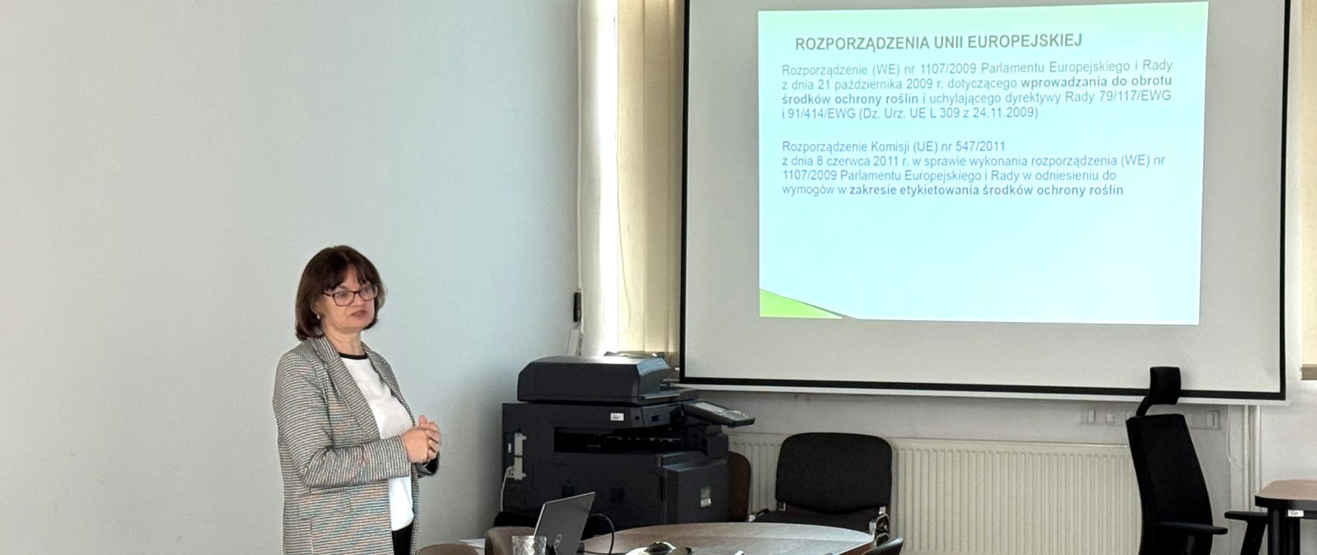 Kobieta ubrana w szarą marynarkę stoi przy stołach na których leży laptop. W tle widać wyświetlaną prezentacje na projektorze. Pomieszczenie jest jasne. 