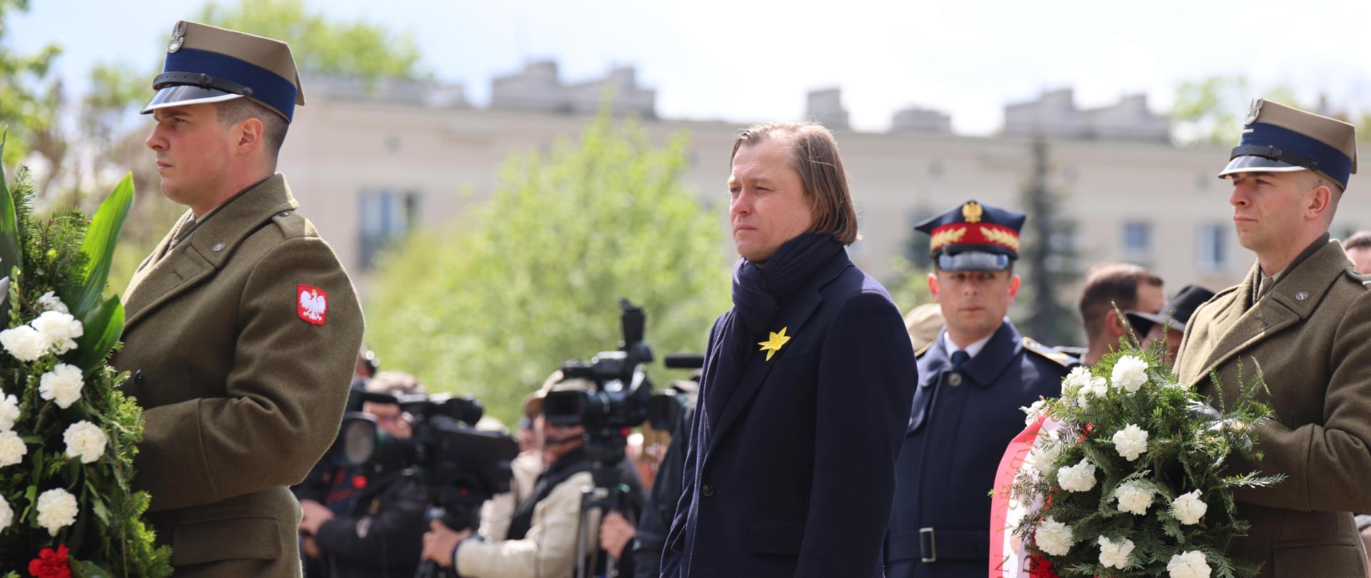 Na zdjęciu mężczyzna. Ma przypięty żółty kwiat do płaszcza. 