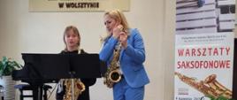 Uczennica wraz z nauczycielką stoją, trzymając saksofony. Patrzą w nuty ustawione na pulpicie.