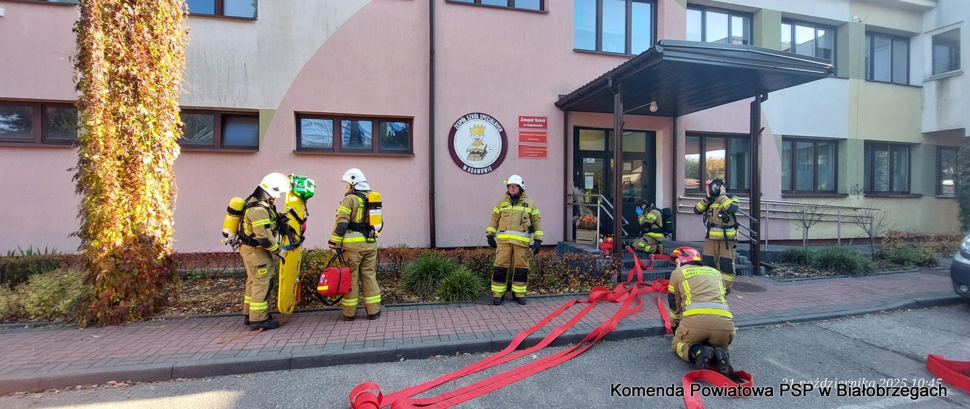 Sześciu strażaków w ubraniach bojowych i hełmach, z których część ma aparaty oddechowe, stoi przed wejściem do różowego budynku szkoły. Dwóch z nich klęczy, rozwijając czerwone węże strażackie na ziemi przed wejściem.