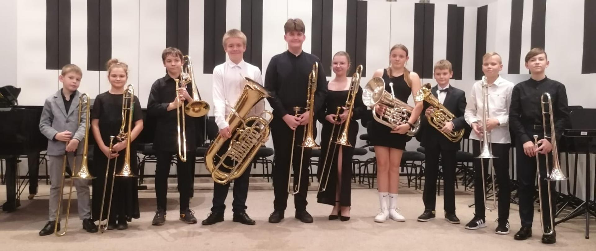 Ośmiu chłopców i dwie dziewczyny stoją w rzędzie na estradzie auli PSM trzymając przed sobą swoje instrumenty.