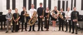 Ośmiu chłopców i dwie dziewczyny stoją w rzędzie na estradzie auli PSM ze swoimi instrumentami.