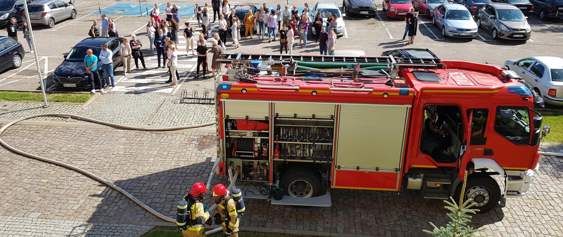 Na pierwszym planie ciężarówka strażacka, przed nią strażacy w sprzęcie przygotowują się do wejścia do wnętrza budynku. W tle stoją zgromadzeni pracownicy i petenci urzędu, którzy po ogłoszeniu alarmu ewakuowali się w bezpieczne miejsce.