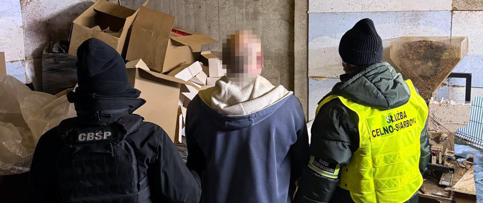 Trzy osoby stoją w magazynie przed stertą kartonów i foliowych opakowań; dwie z osób mają na odzieży widoczne oznaczenia służb.