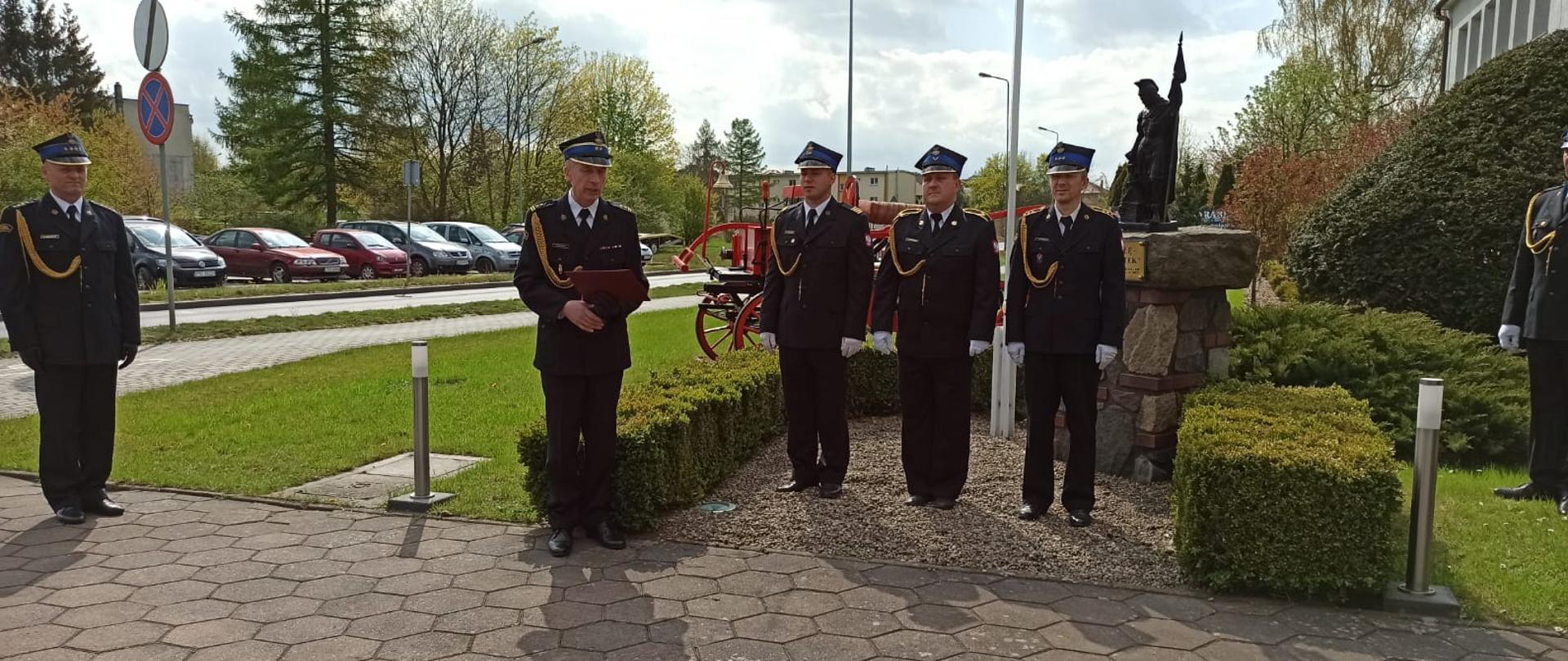 Zdjęcie przedstawia poczet flagowy składający się z trzech strażaków w mundurach galowych, Komendanta Powiatowego czytającego życzenia z okazji dnia strażaka oraz dowódcy uroczystości. W tle znajduje się czerwona sikawka konna.
