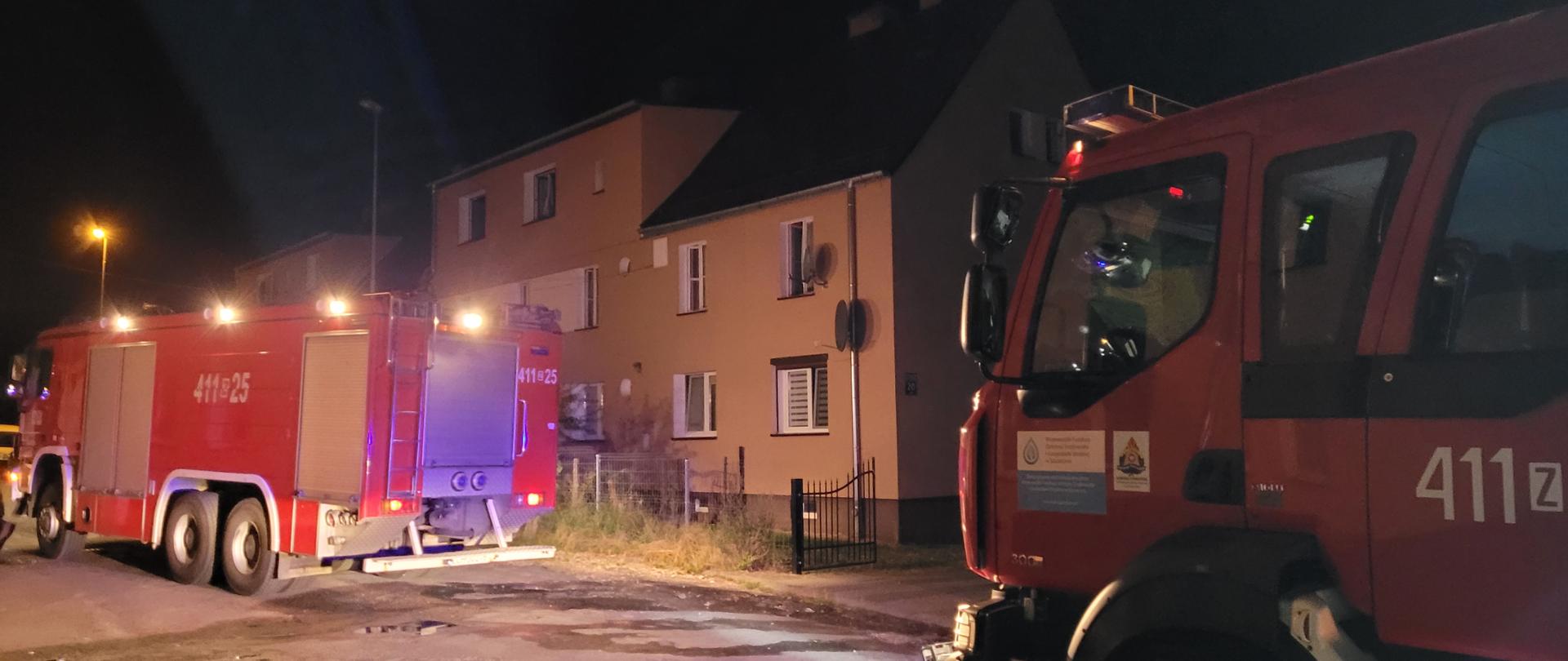 Na fotografii znajdują się dwa wozy strażackie zaparkowane przy drodze. Oba pojazdy mają włączone sygnały świetlne. Po prawej stronie pojazdów znajdują się dwa budynki mieszkalne. Zdjęcie wykonano w porze nocnej,